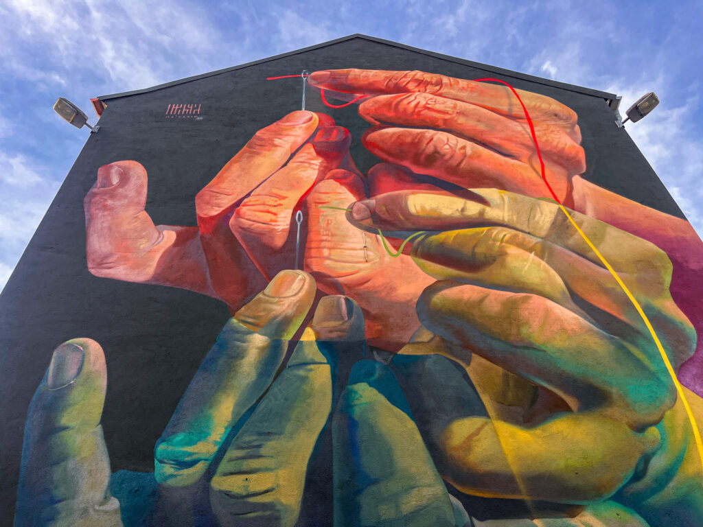 The Needle's Eye – Mural von Case Ma’Claim in Aschaffenburg zeigt Hände, die einen Faden durch ein Nadelöhr führen.