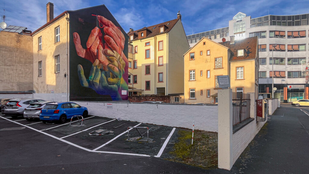 The Needle's Eye – Mural von Case Ma’Claim in Aschaffenburg zeigt Hände, die einen Faden durch ein Nadelöhr führen.