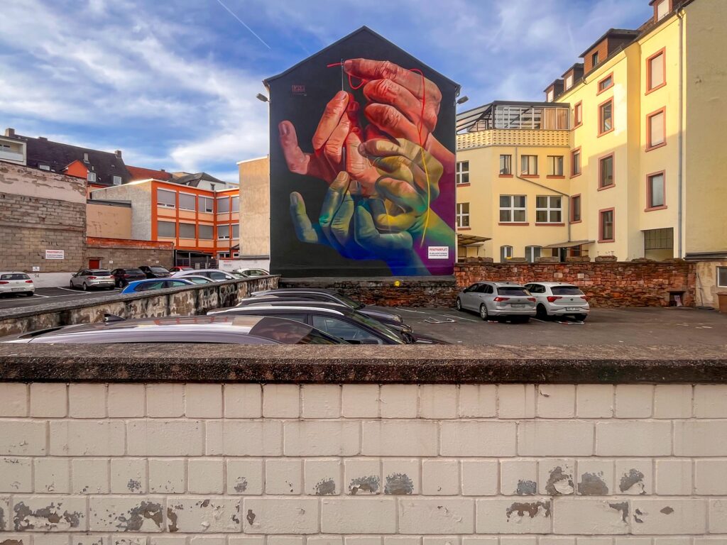 The Needle's Eye – Mural von Case Ma’Claim in Aschaffenburg zeigt Hände, die einen Faden durch ein Nadelöhr führen.