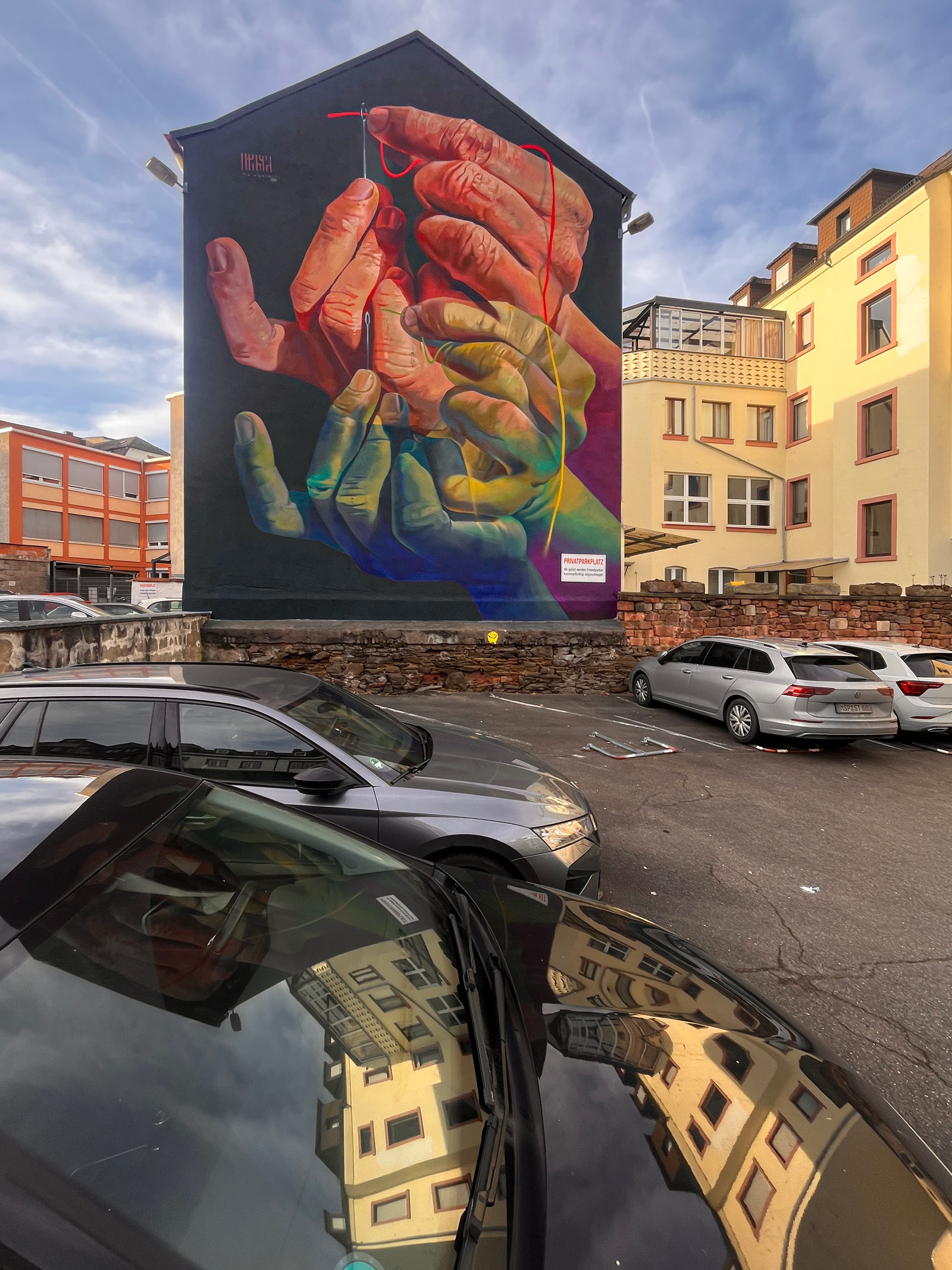 The Needle's Eye – Mural von Case Ma’Claim in Aschaffenburg zeigt Hände, die einen Faden durch ein Nadelöhr führen.
