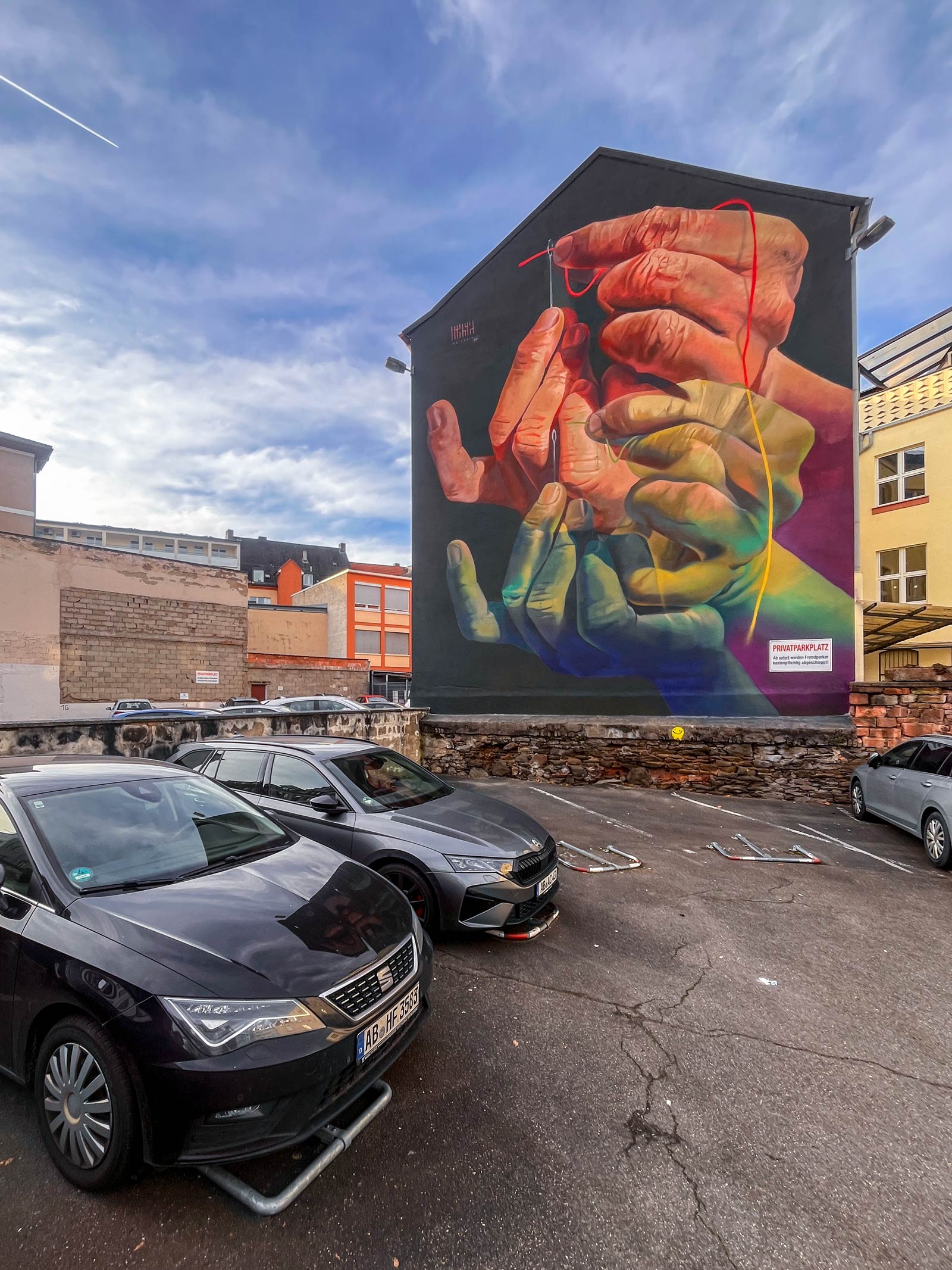 The Needle's Eye – Mural von Case Ma’Claim in Aschaffenburg zeigt Hände, die einen Faden durch ein Nadelöhr führen.