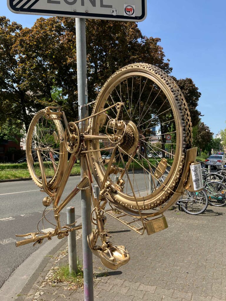 Fahrrad zuerst pink dann gold, hochkant befestigt an Verkehrsschild-Pfosten in Frankfurt
