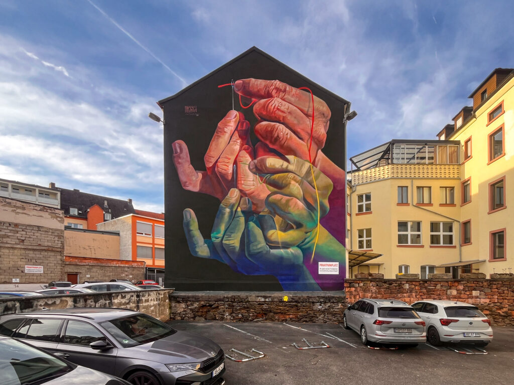 The Needle's Eye – Mural von Case Ma’Claim in Aschaffenburg zeigt Hände, die einen Faden durch ein Nadelöhr führen.